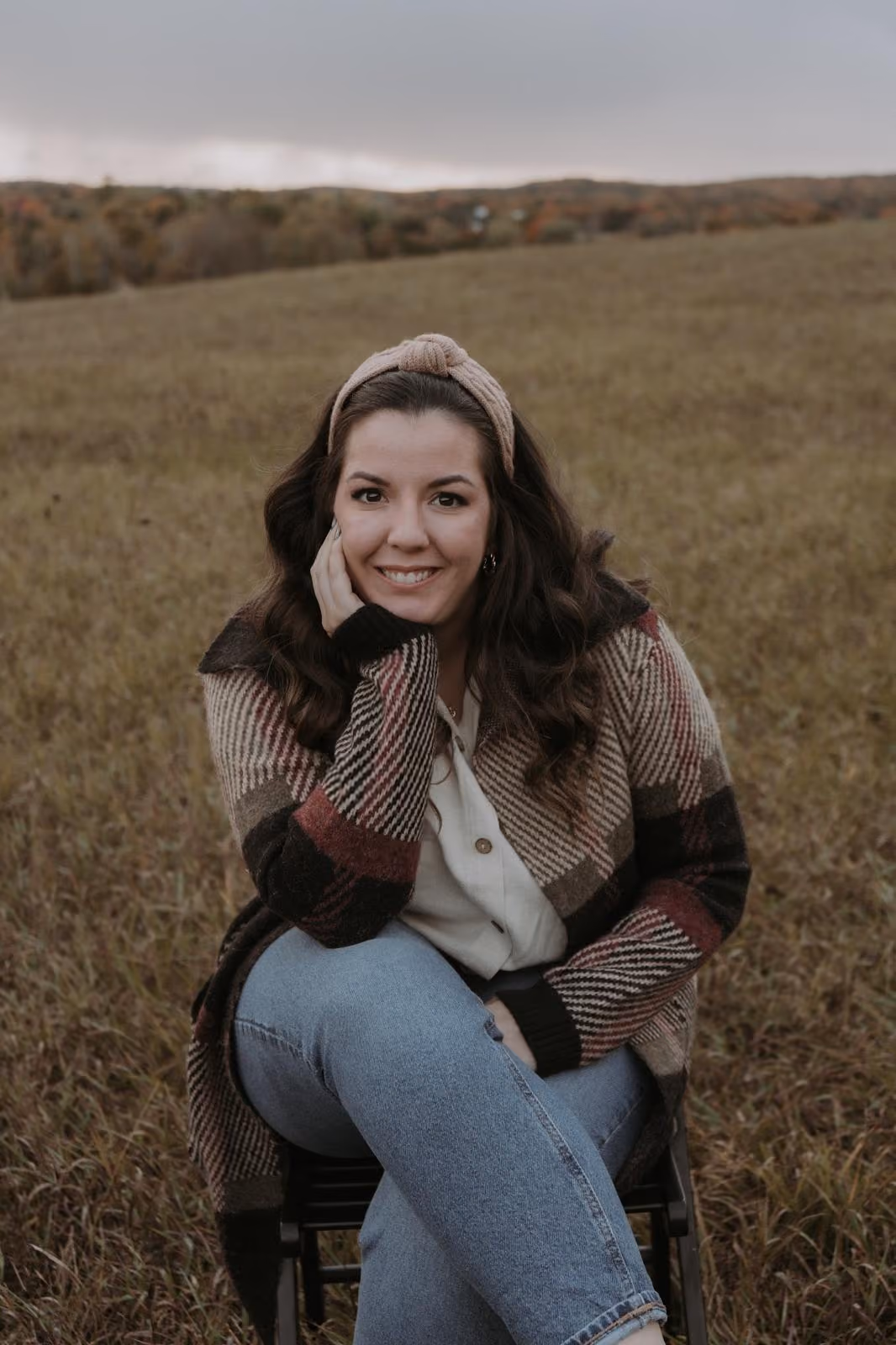 Kate Duquette seated outdoors in an autumn field, resting her chin on her hand and smiling warmly.